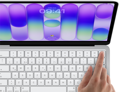 MacBook Neo, gümüş rengi, ekranda oturum açma penceresi gösteriliyor, kullanıcının parmağı klavyenin sağ üst köşesindeki Touch ID sensörüne dokunuyor